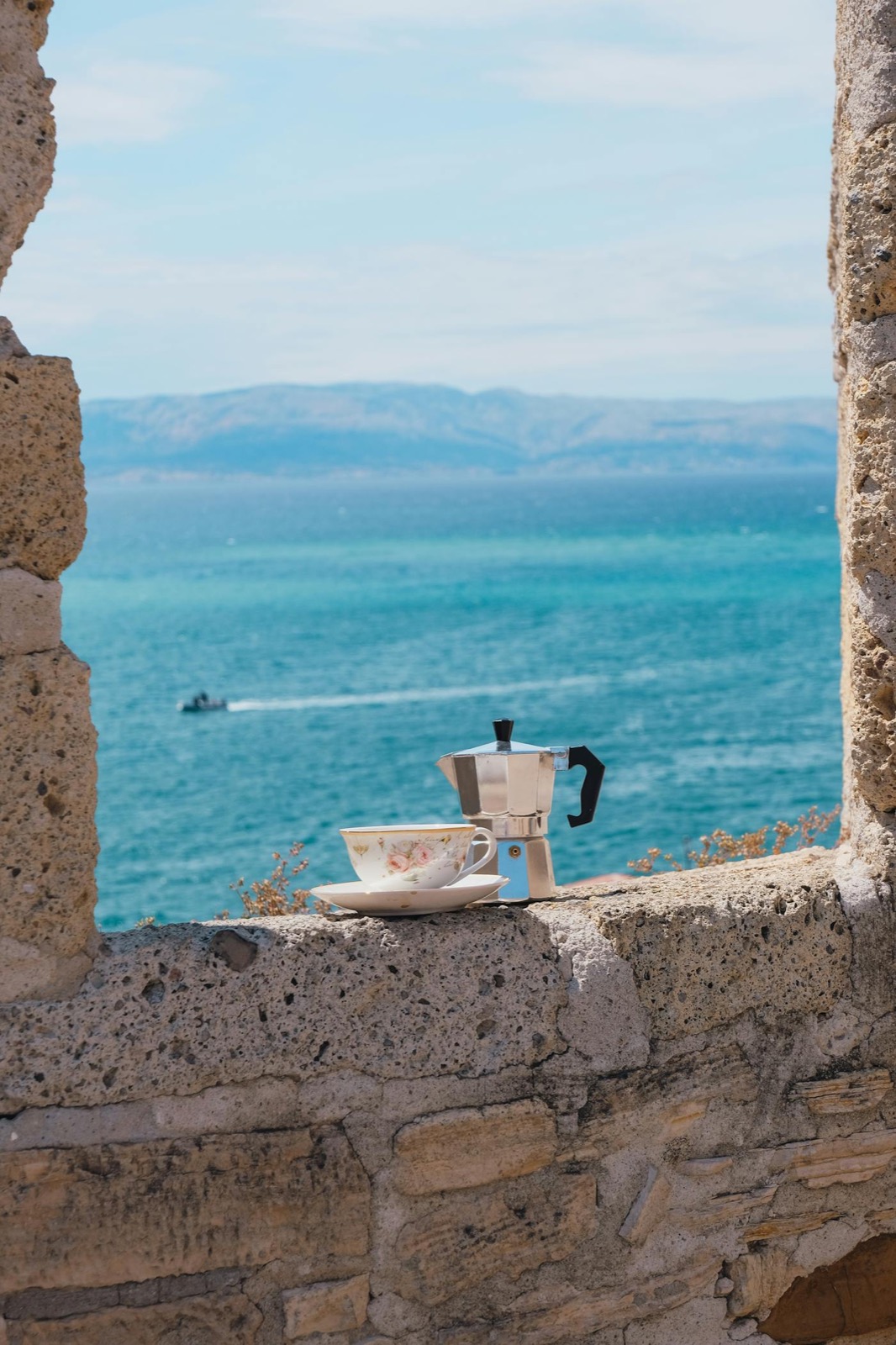 Cafetière italienne moka et tasse posées sur un mur de pierre méditerranéen, mer turquoise en arrière-plan
