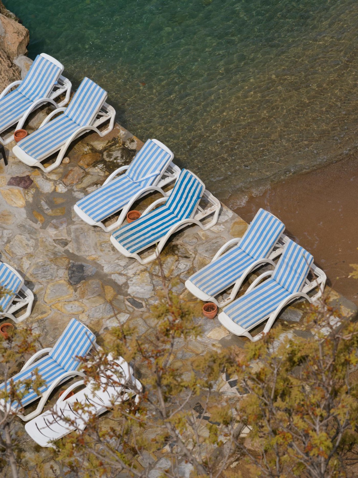 Vue aérienne de transats à rayures bleues et blanches sur un terrasse de pierre face à la mer turquoise